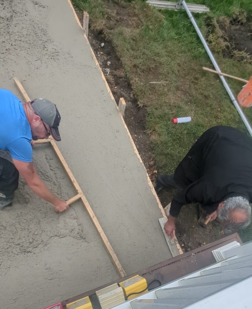 Two Sarnia concrete driveway contractors are screeding and edging a freshly poured concrete driveway for a Calmachie house on Egermont Rd, with staked forms and tools visible.